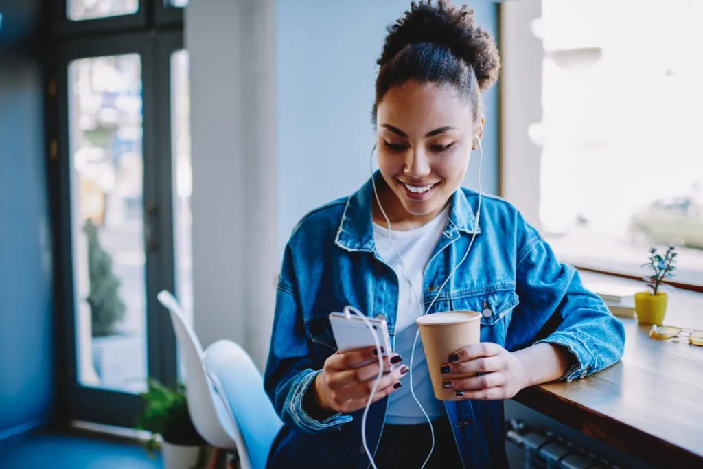 Happy female meloman typing name of song for search and download from multimedia application on cellular phone during coffee time in cafeteria,positive hipster girl listening audio book via headphones